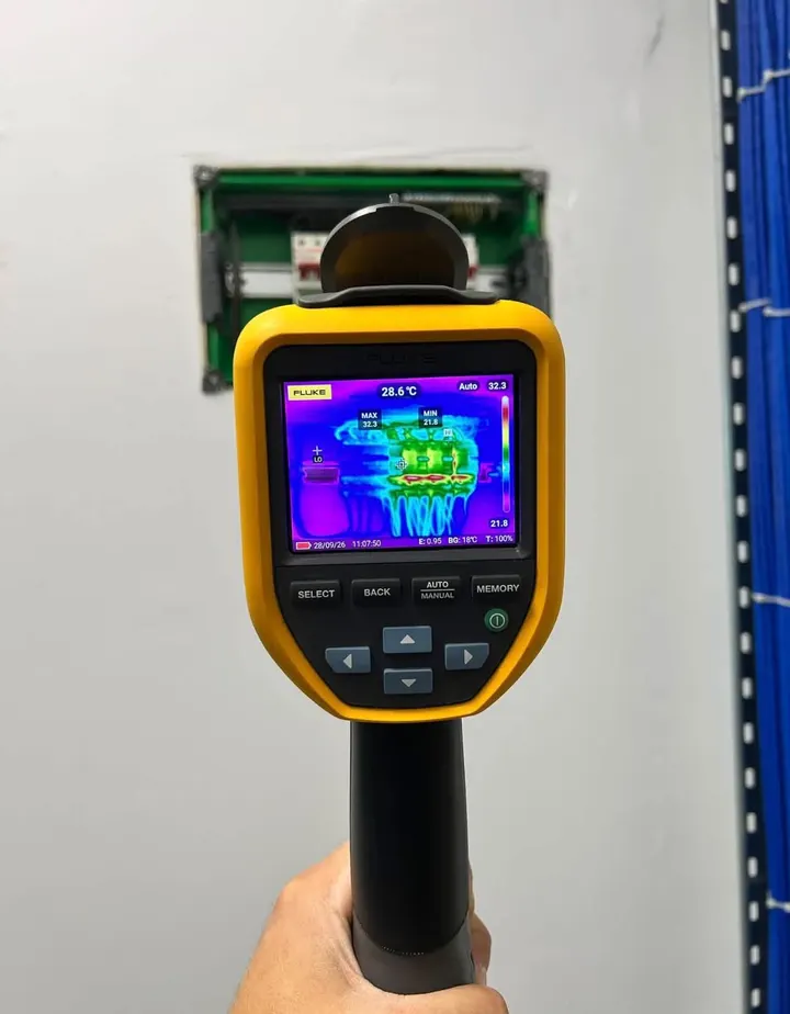 Cameră cu termoviziune ținută în fața unui tablou electric, cu imaginea termică pe ecran